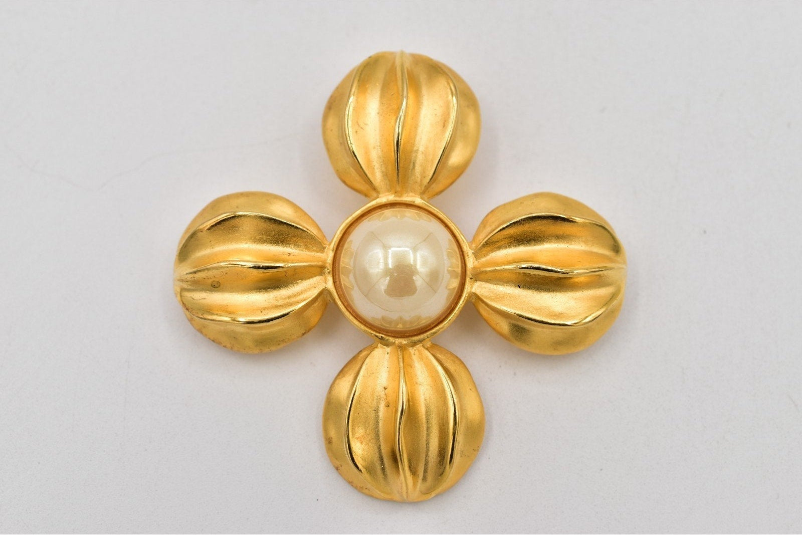 A vintage-style gold flower-shaped brooch with a central white pearl, photographed on a plain light gray background.