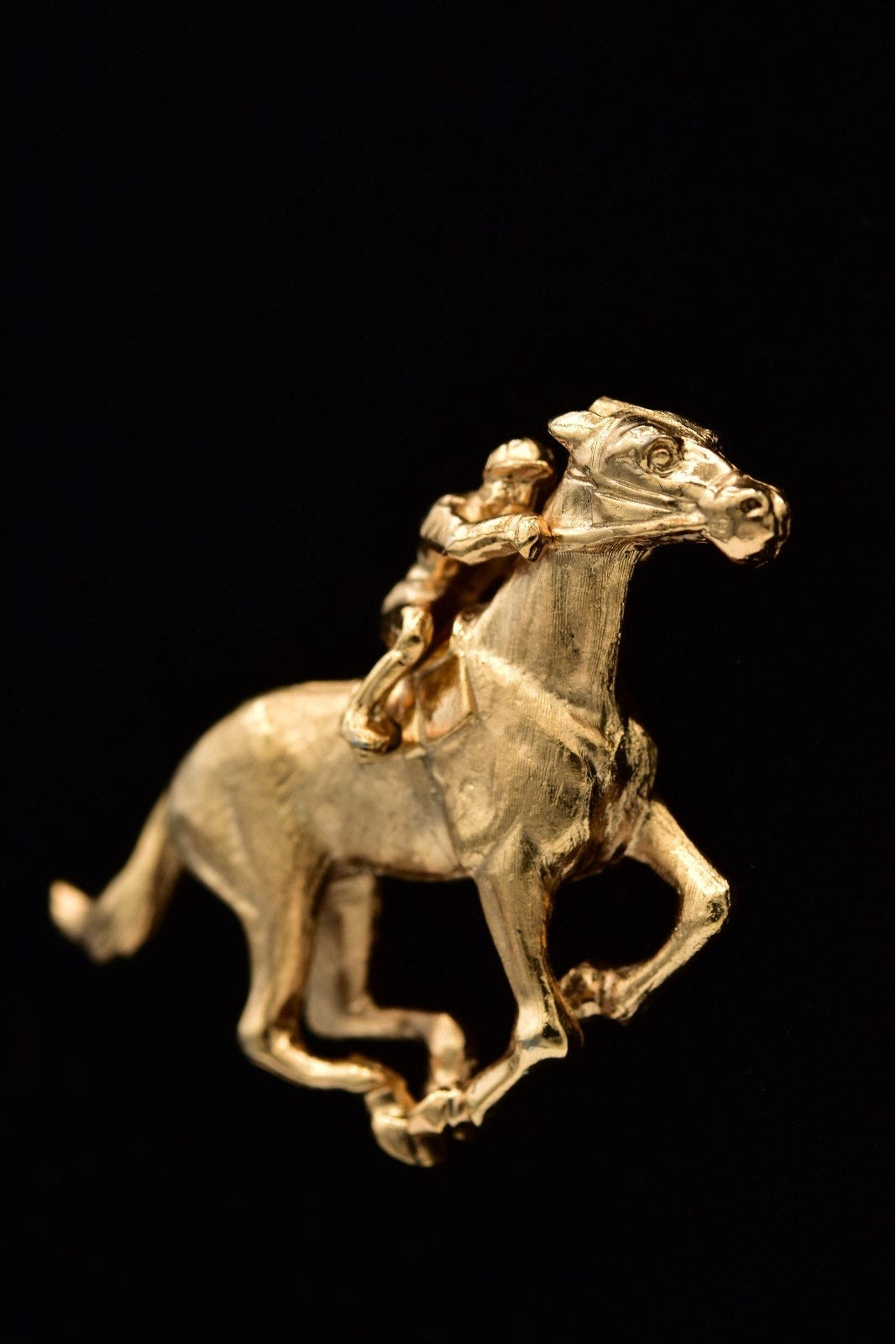 A small gold equestrian figurine or charm depicting a horse with a rider, shown against a solid black background.