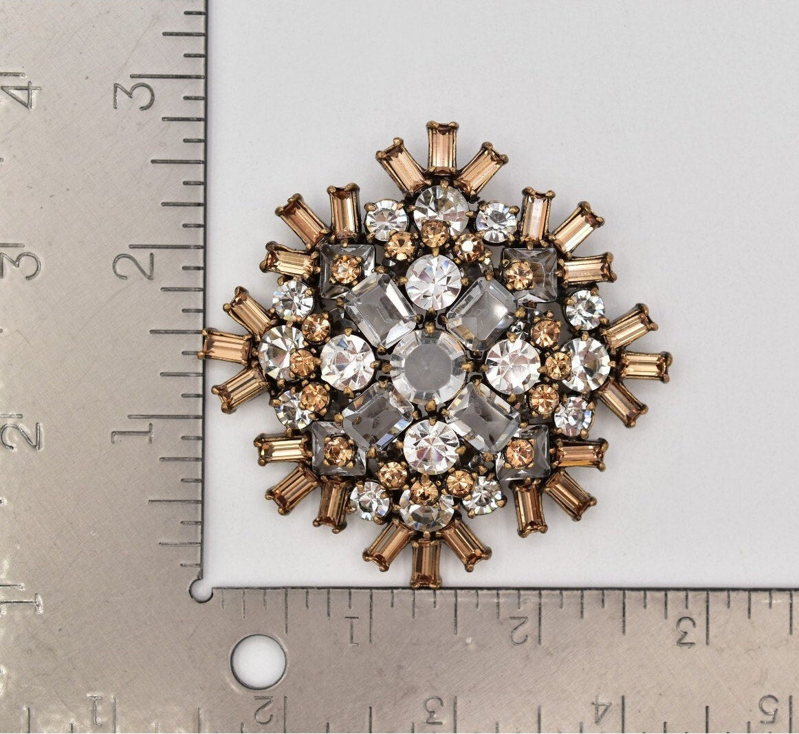 Decorative brooch with crystals on a white background, measured against a ruler.
