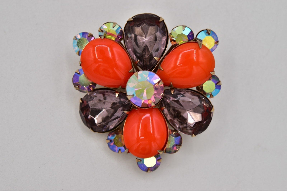 A vintage-style floral brooch featuring four bright orange-red oval stones, four dark purple teardrop stones, and a central iridescent round crystal, surrounded by small iridescent round rhinestones on a light gray background.