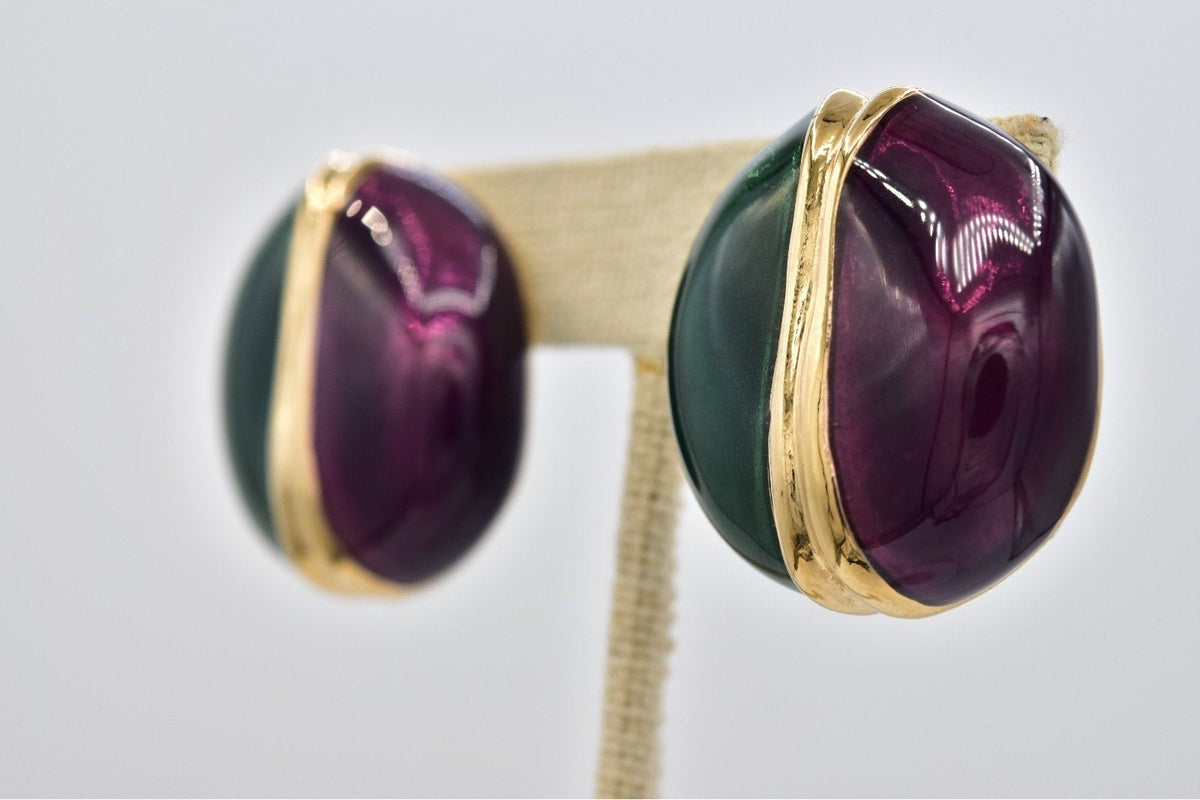 A pair of oval stud earrings with deep purple cabochon stones and dark green enamel, set in gold-tone metal, displayed on a cream-colored jewelry stand against a light gray background.