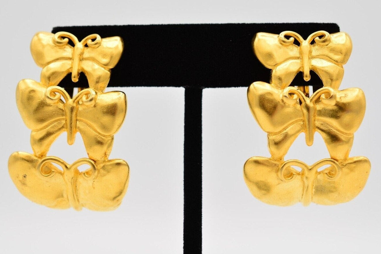 A pair of gold butterfly-shaped drop earrings displayed on a black T-stand against a light gray background.