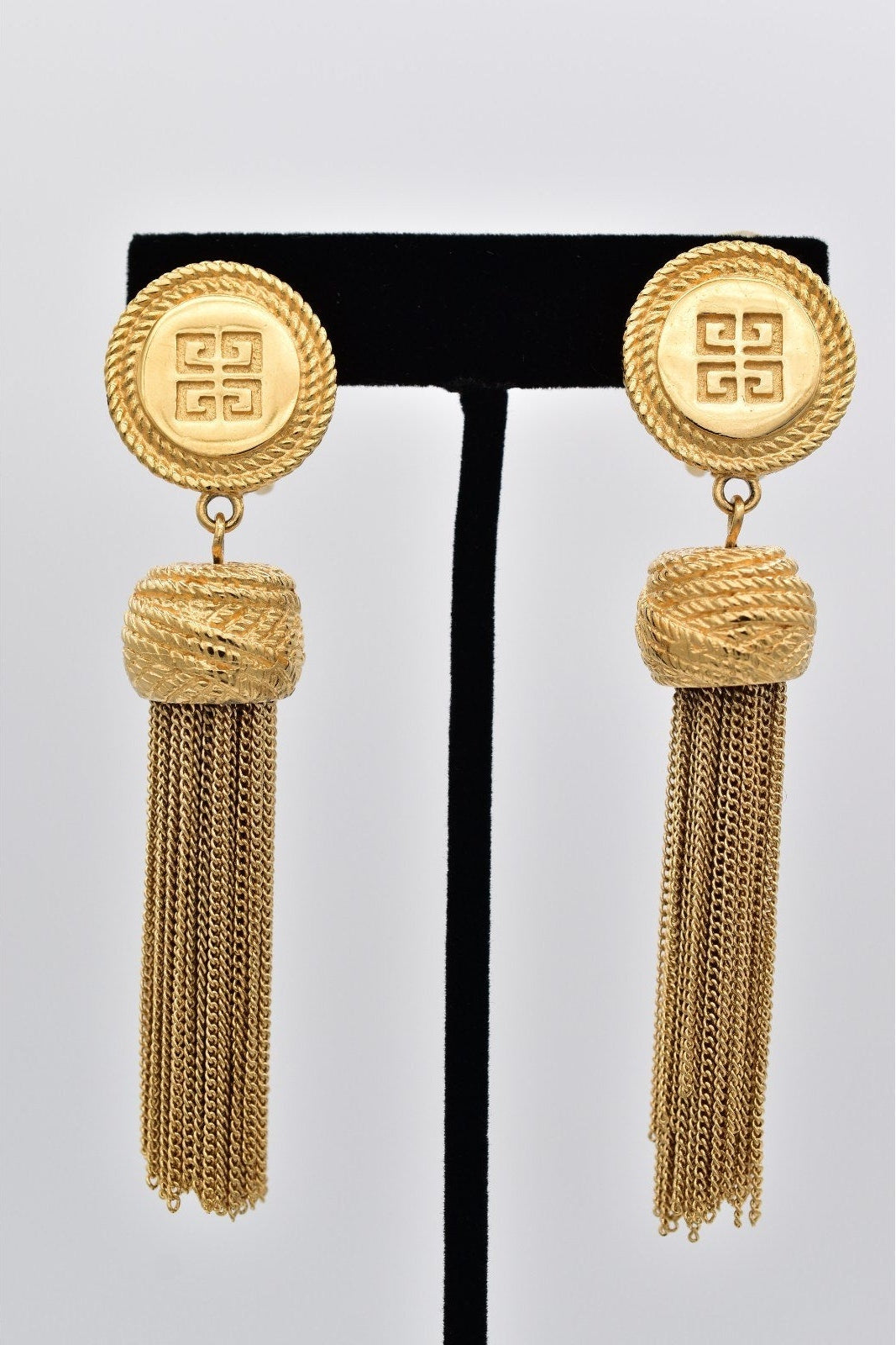 Gold tassel earrings on a black stand against a light gray background