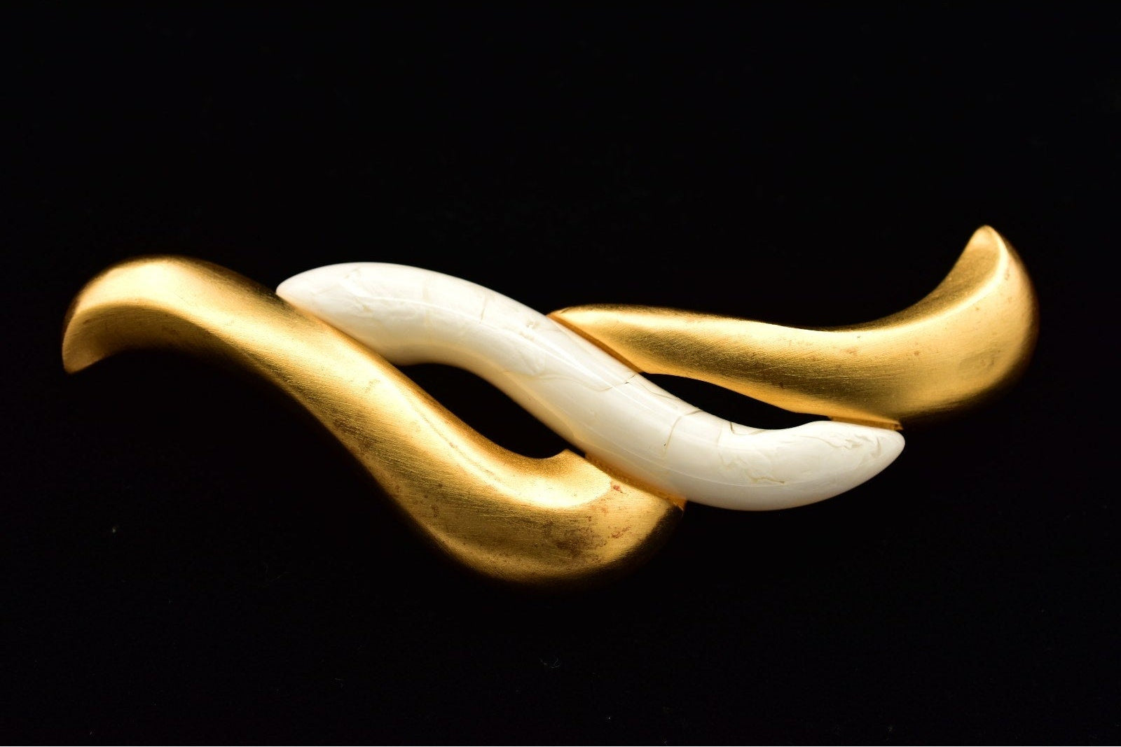 A modern abstract wavy knot brooch featuring polished gold and white curved segments, presented on a solid black background.