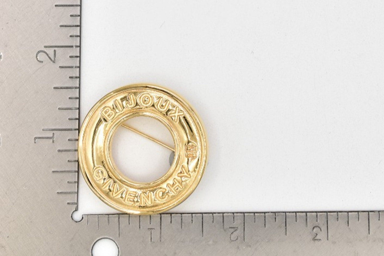 A small round gold-tone metal pin or charm with a circular cutout and engraved text, shown on a white background with ruler edges visible for scale.