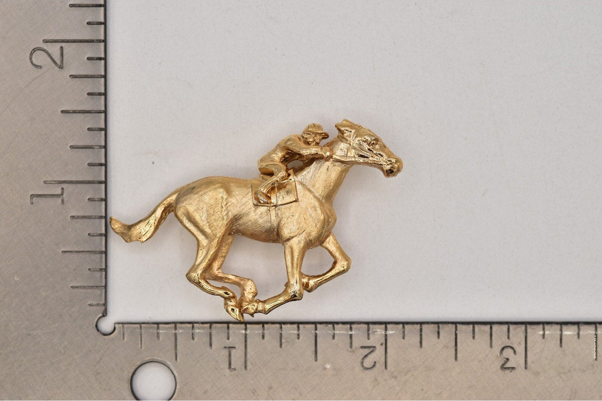 A small gold-colored horse and jockey figurine, shown next to a metal ruler for scale on a light gray background.