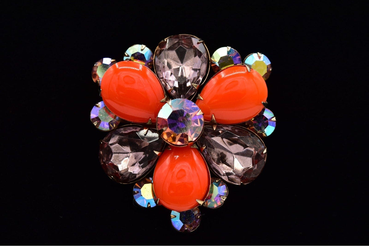 A vintage-style floral brooch featuring four bright orange cabochon stones surrounded by faceted taupe-gray and iridescent rhinestones on a black background.