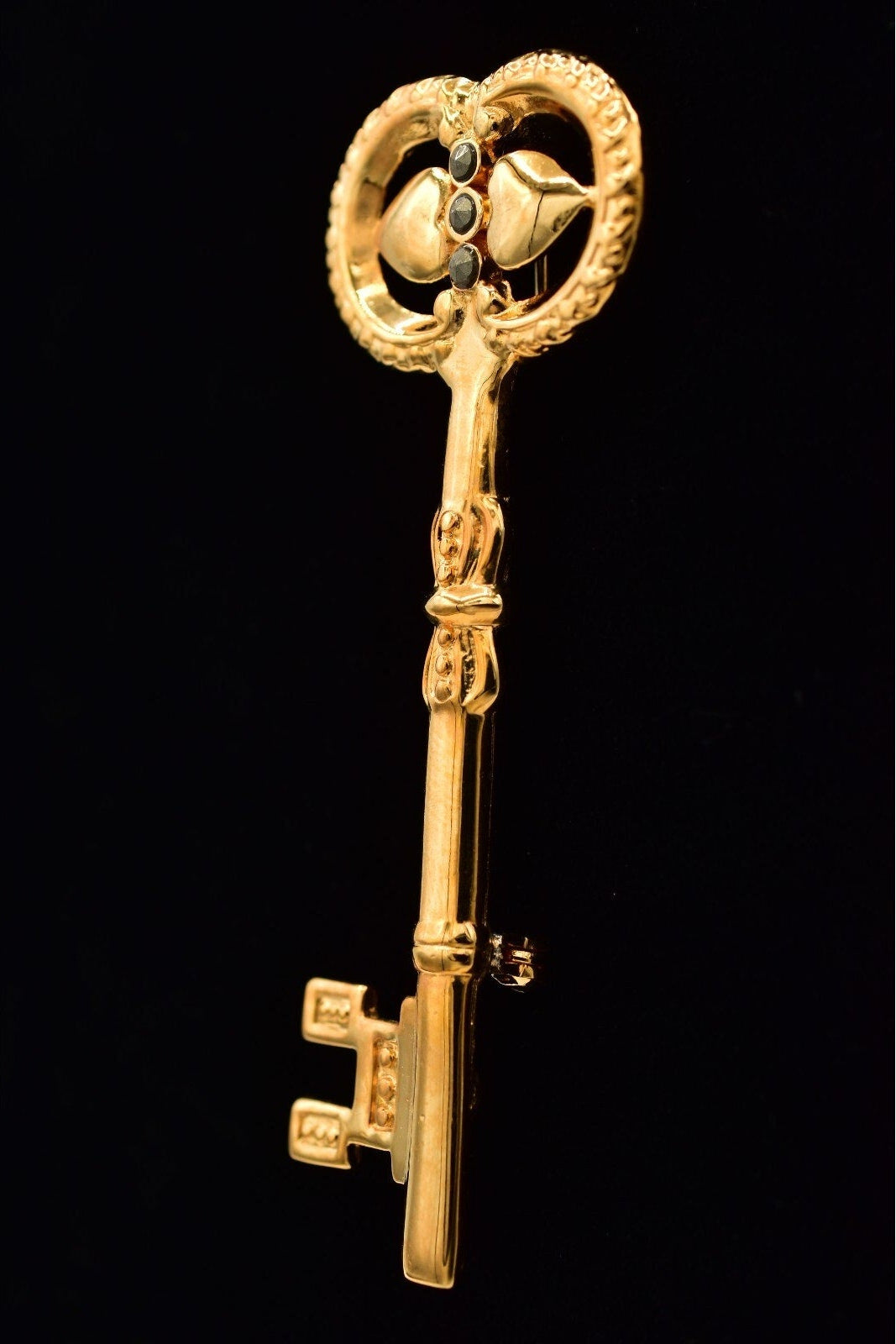 A vintage-style gold key brooch or pendant with ornate bird and floral details on the bow, shown against a solid black background.