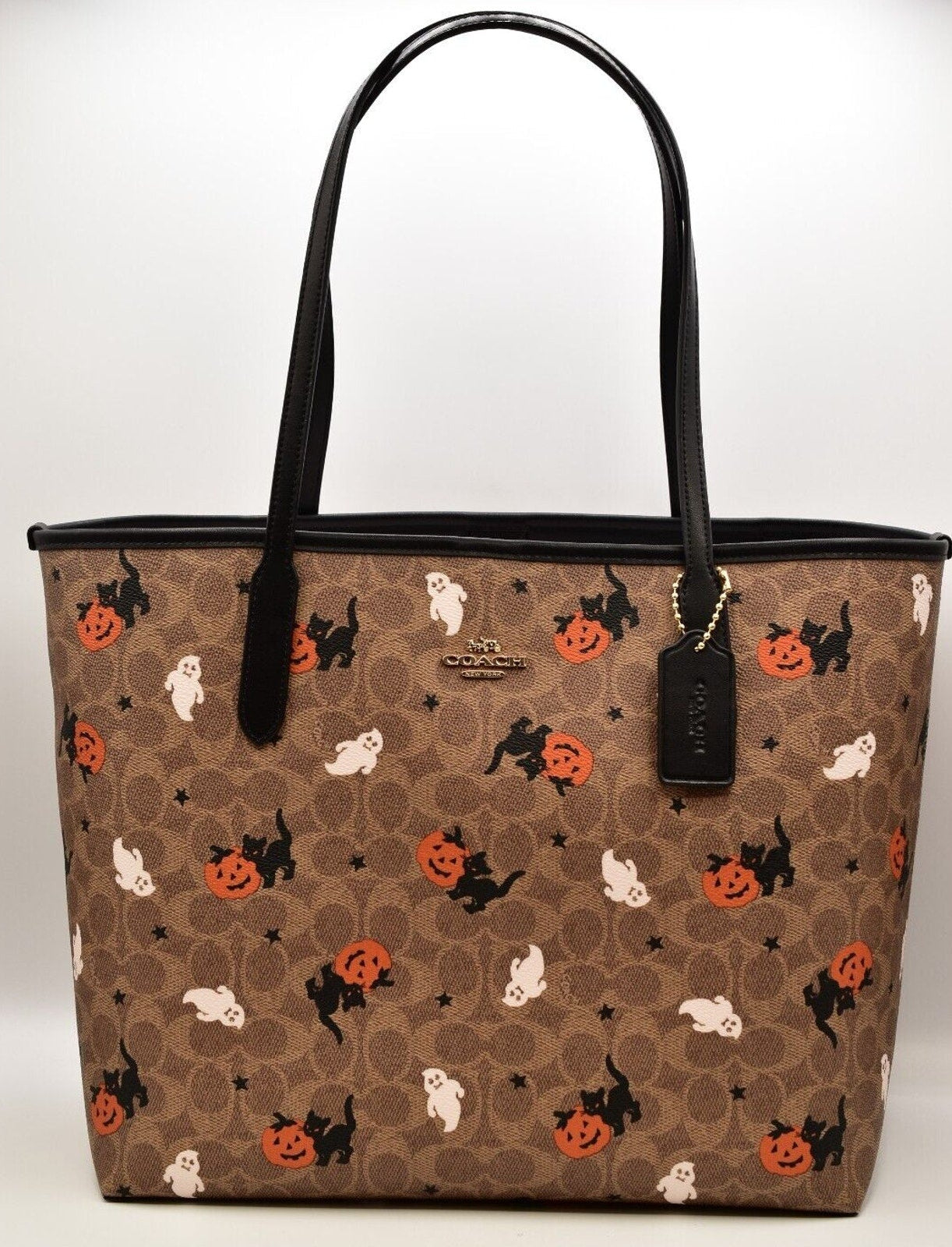 A brown Coach tote bag featuring a Halloween-themed monogram pattern with black cats, orange pumpkins, and white ghosts, complete with black leather handles and a hanging brand tag.