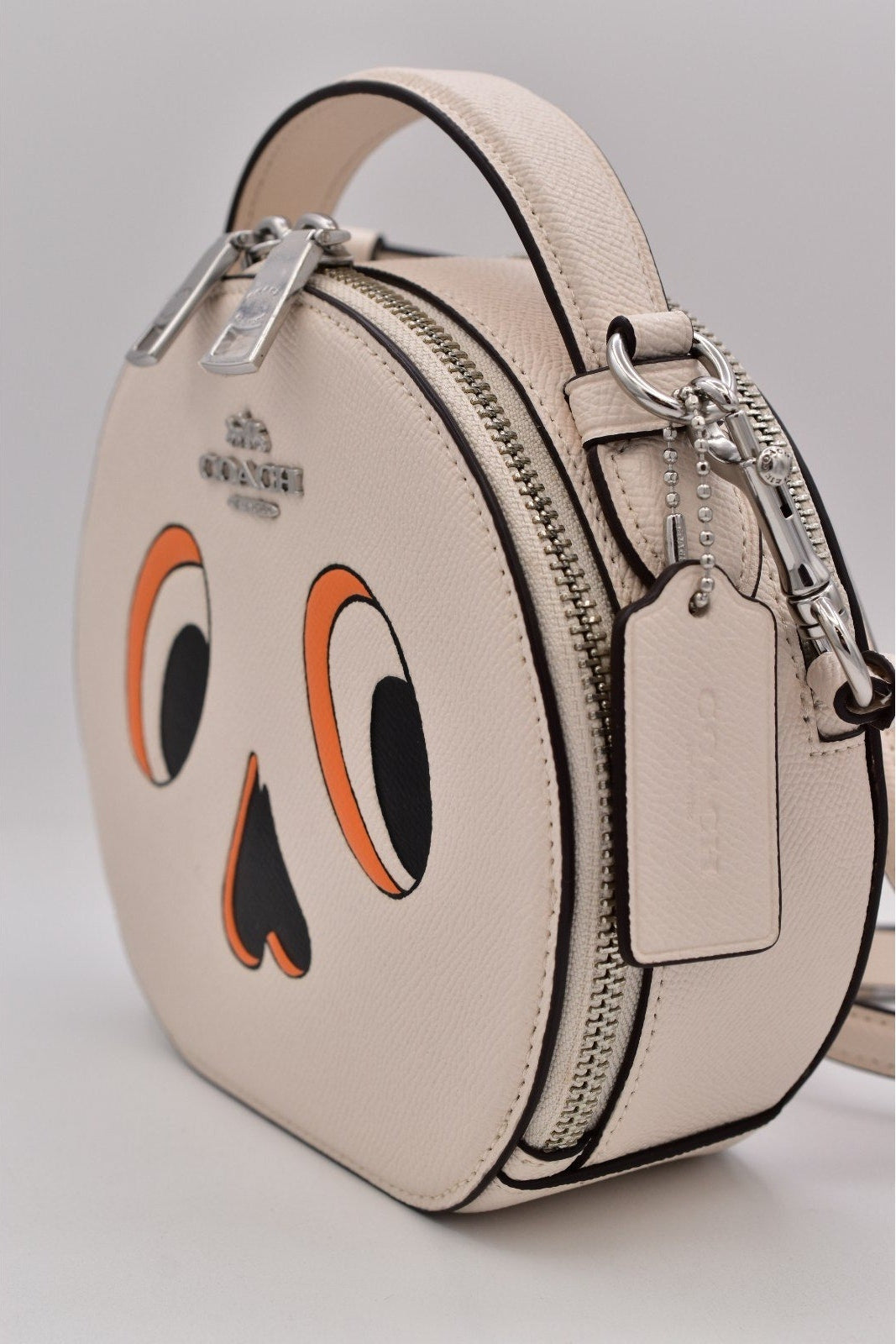 A Coach crossbody bag in light beige leather with a playful cartoon face design featuring orange and black eyes and a nose, shown on a neutral gray background.