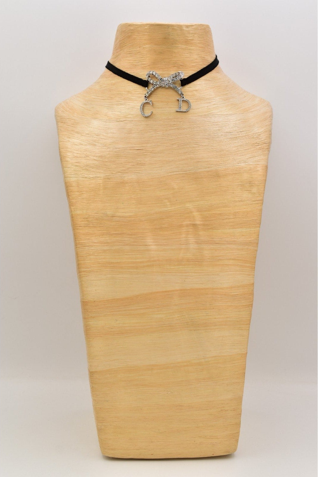 A black cord choker necklace with a silver bow charm and dangling 'C' and 'D' charms, displayed on a light wood neck mannequin against a neutral light gray background.
