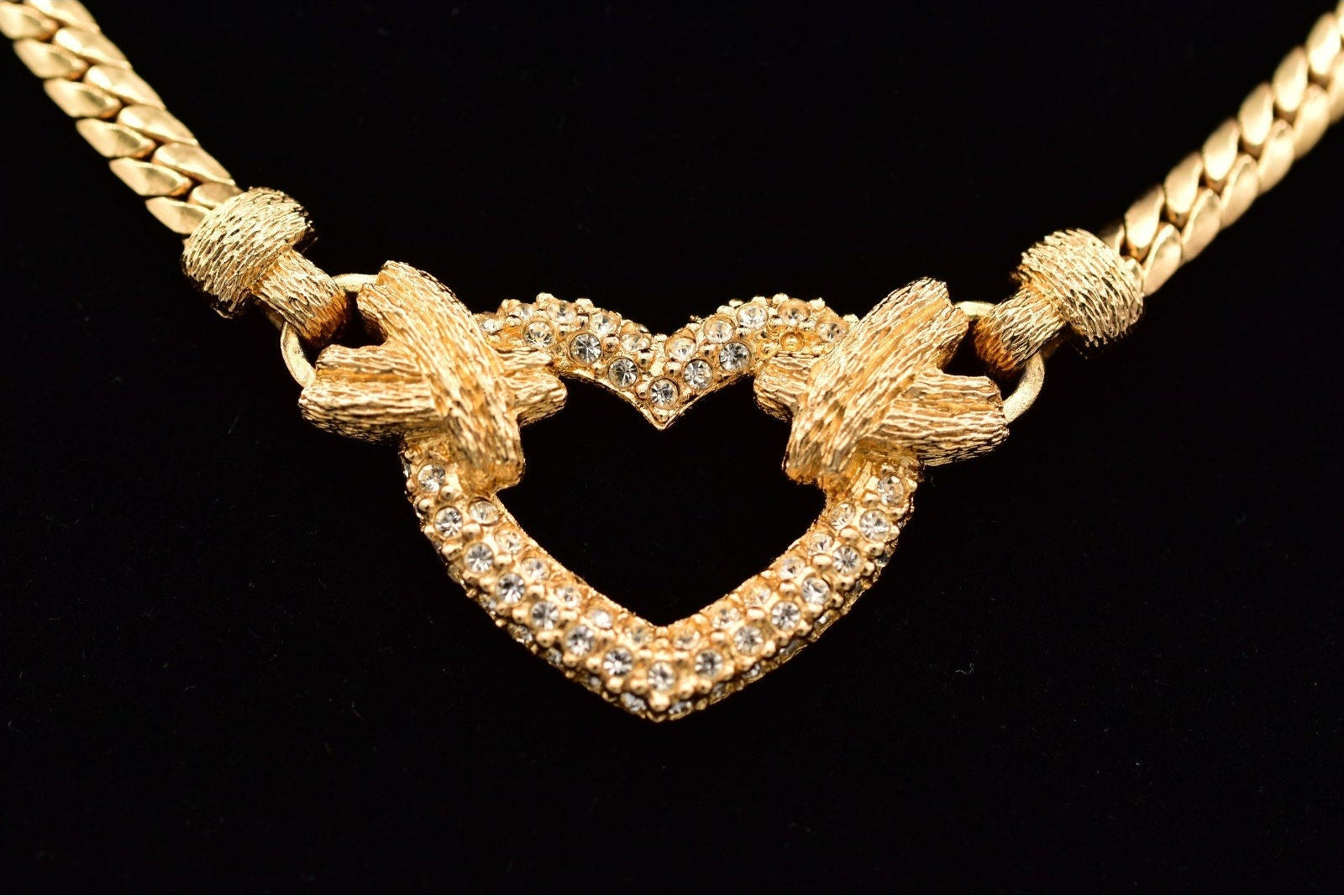 A gold heart pendant necklace with a textured rope-knot design at the top and clear pavé stones along the bottom curve, displayed on a black background.