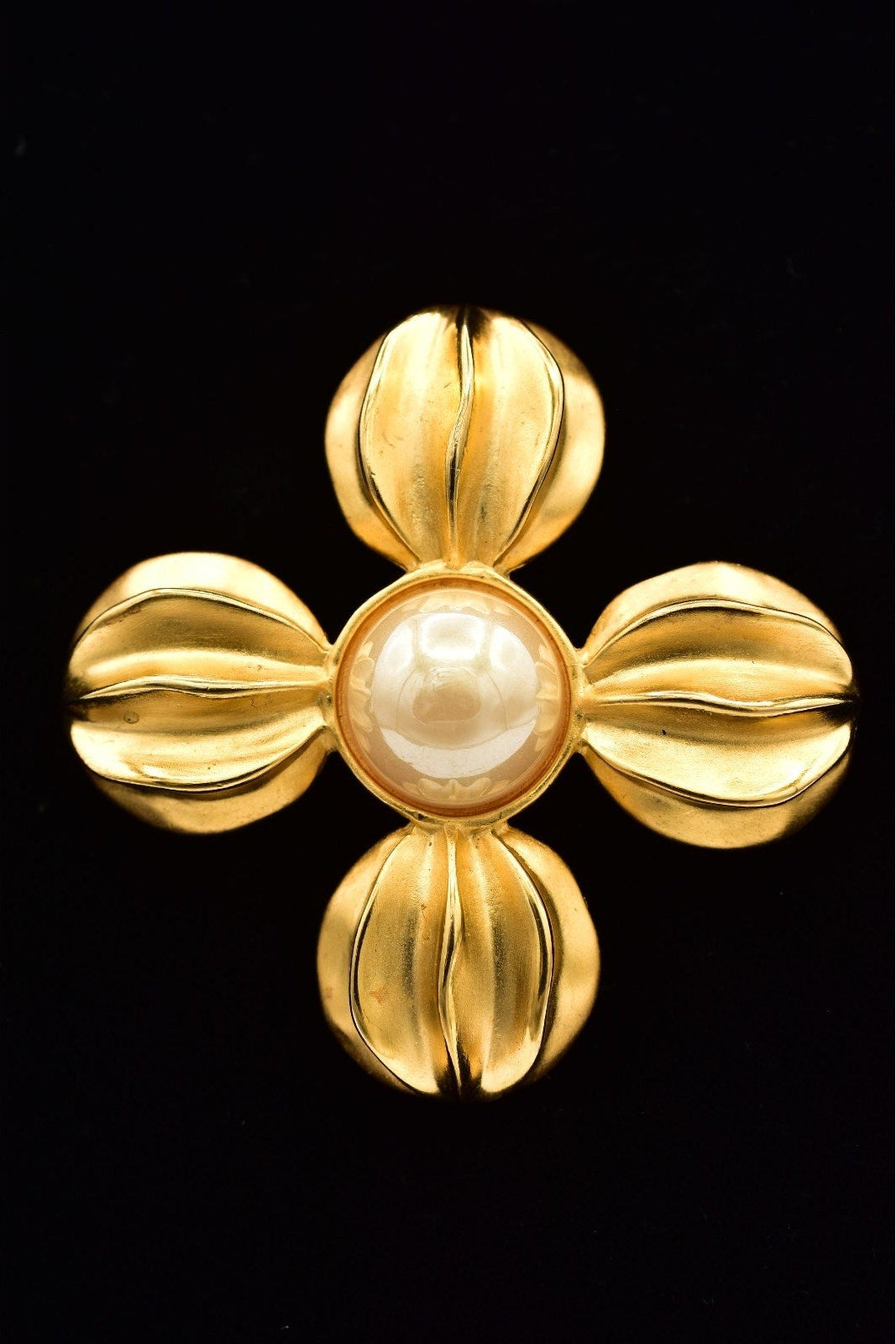 A vintage-style gold cross brooch featuring a central round white pearl surrounded by four textured, petal-like gold arms, set against a solid black background.