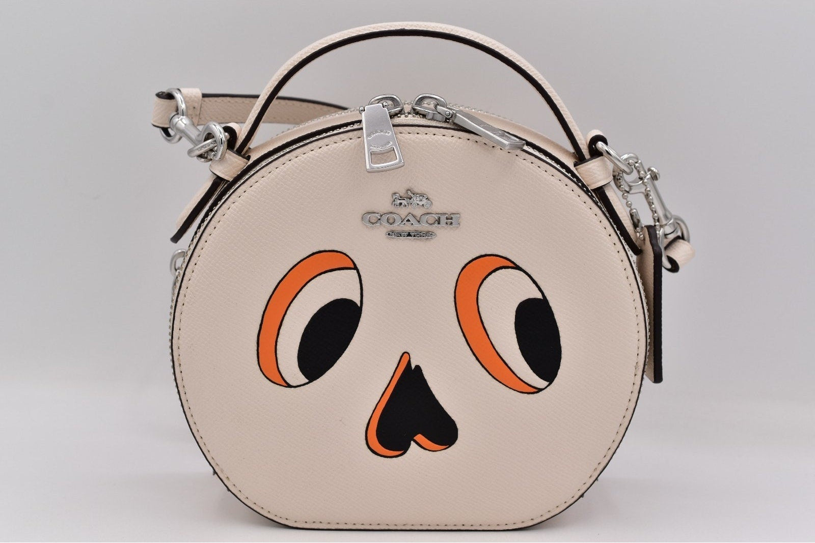 A round Coach handbag in light beige leather with a cartoon face design featuring orange and black eyes and a nose, complete with a top handle and silver-tone hardware.