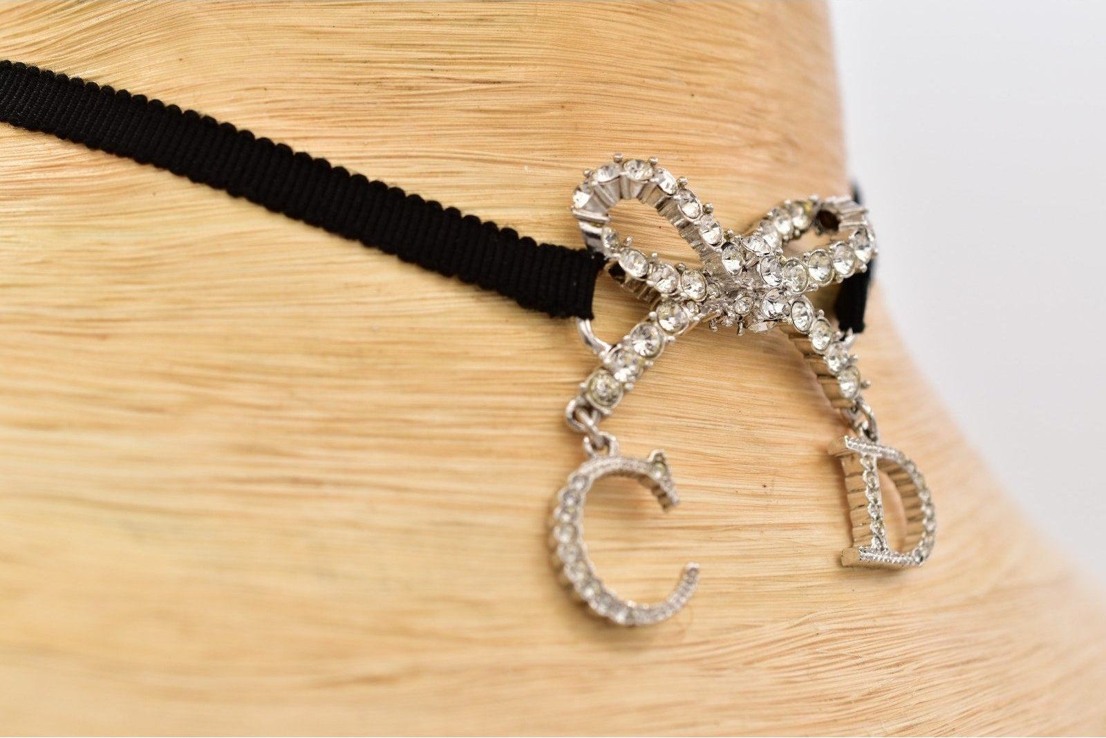 A close-up of a black ribbon choker necklace with a silver bow charm encrusted with clear rhinestones, featuring dangling letter charms 'C' and 'D', displayed on a light wood mannequin stand.
