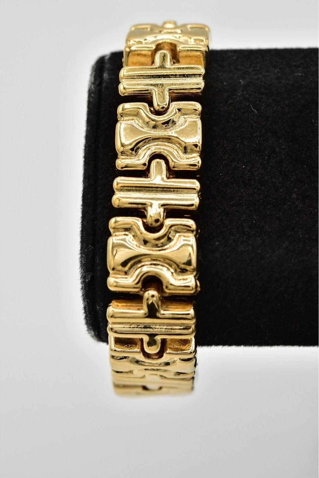 A gold-tone metal link bracelet with a distinctive wavy, interlocking geometric design, displayed on a black velvet jewelry stand against a light gray background.