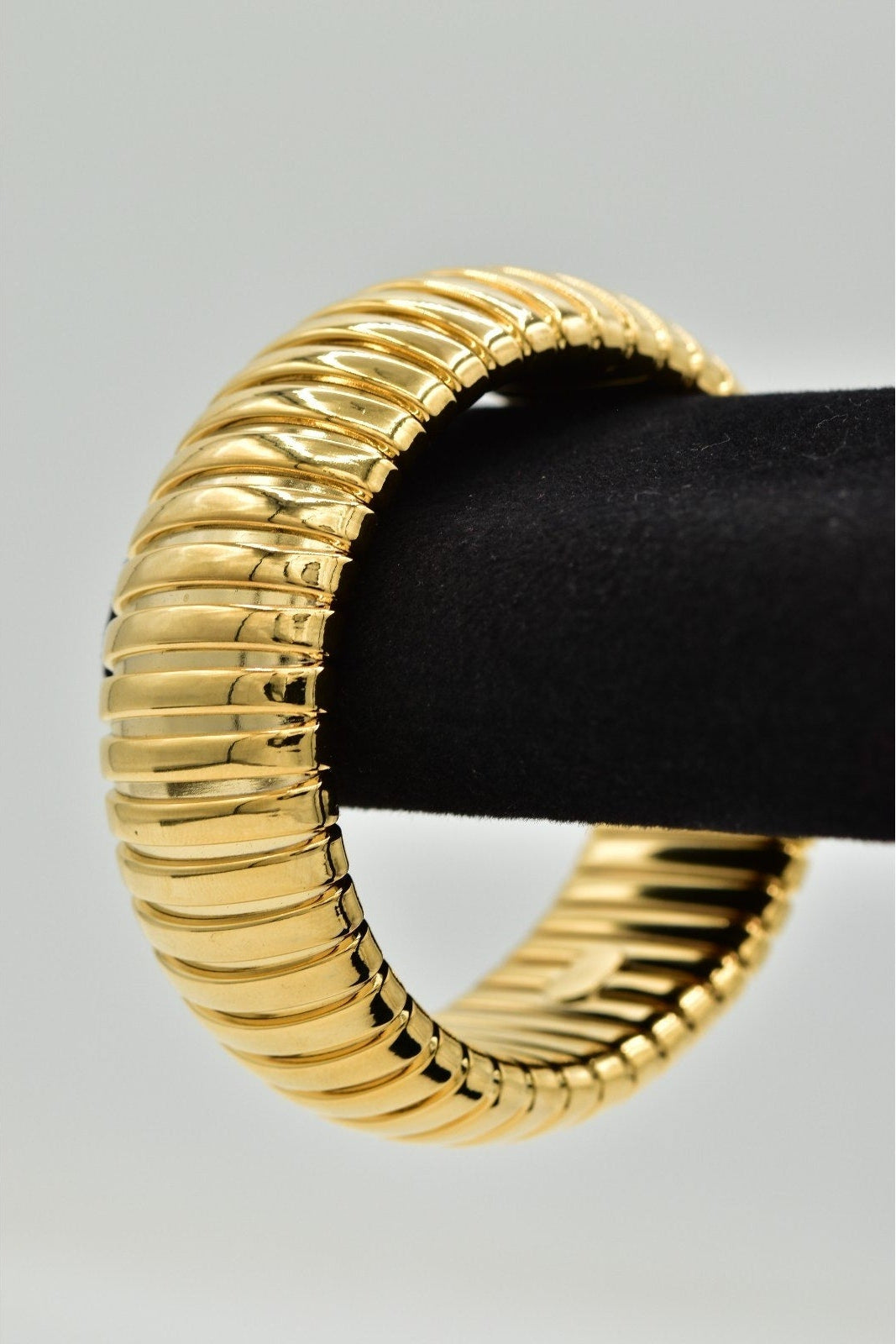 A wide, chunky gold bangle bracelet with a ribbed, snake-like textured pattern, displayed on a black velvet ring stand against a light gray background.