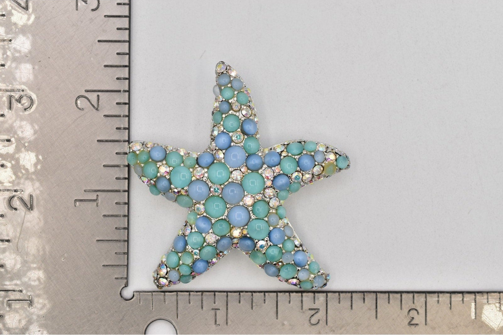 A starfish-shaped jewelry charm or brooch covered in small blue and turquoise rhinestones, shown next to a metal ruler for scale on a light gray background.