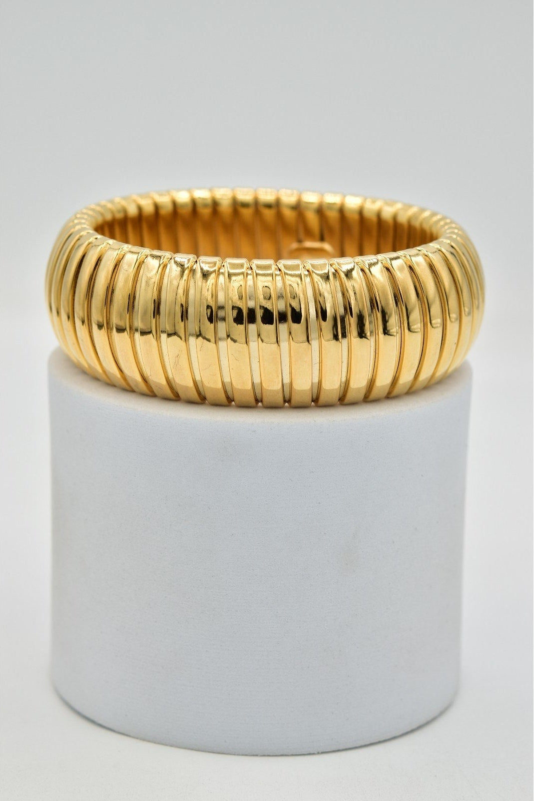A wide, chunky gold bangle bracelet with a ribbed, snake-like textured pattern, displayed on a white cylindrical pedestal against a light gray background.
