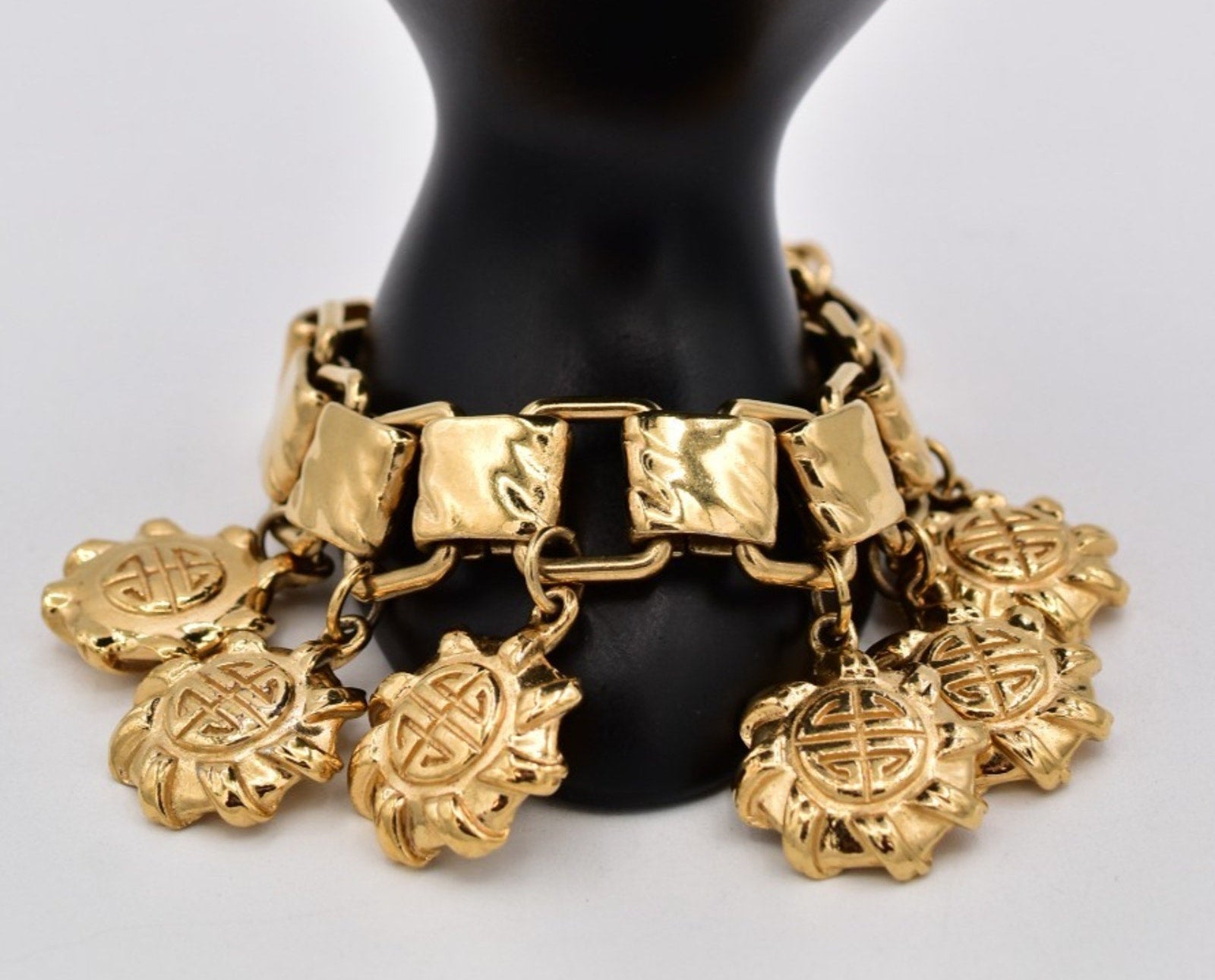 A gold-tone charm bracelet displayed on a black mannequin bust against a light gray background. The bracelet features a textured link chain with multiple dangling lotus-shaped charms, each adorned with a Chinese-style lucky symbol.