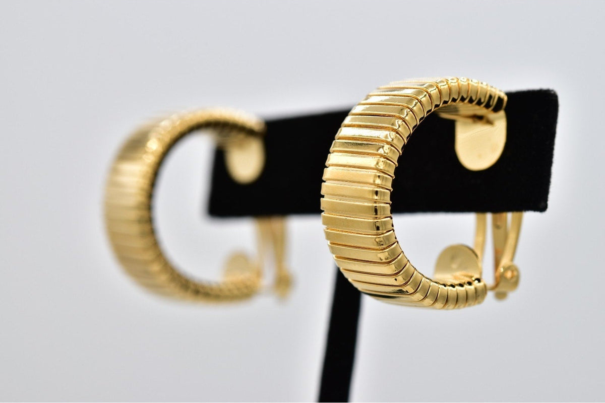 A pair of gold hoop earrings with a textured, ribbed outer band and a smooth inner band, displayed on a black stand against a light gray background.