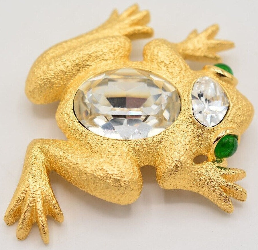 A vintage Christian Dior frog brooch with a golden finish, vibrant green cabochons for eyes, and sparkling crystals on the body.