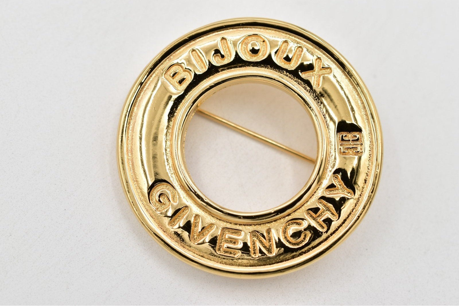 A gold-tone circular Givenchy Bijoux pin with the brand name engraved on the front, set against a plain light background.