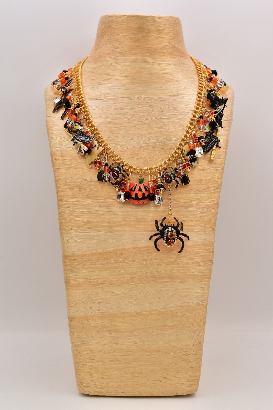 A gold-toned statement necklace with Halloween-themed charms, including spiders, bats, and orange beads, displayed on a light wood mannequin bust.