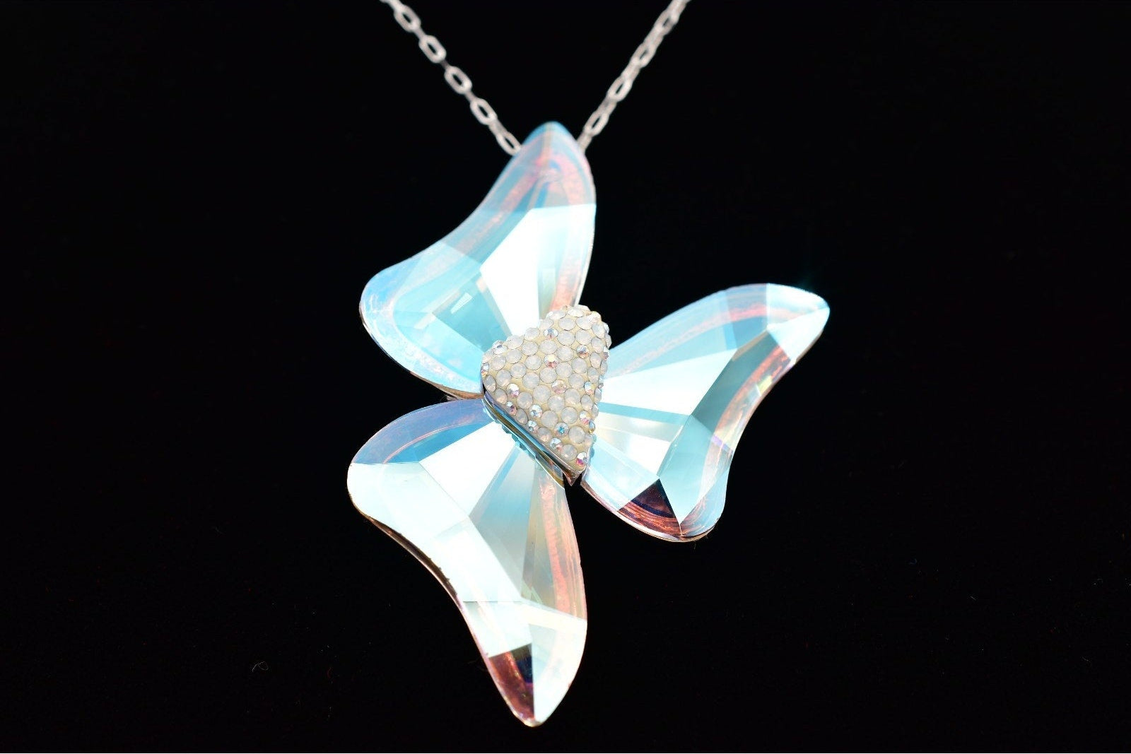 A butterfly-shaped pendant necklace made of iridescent light blue glass with a small heart-shaped cluster of clear rhinestones in the center, hanging from a silver-toned chain against a solid black background.
