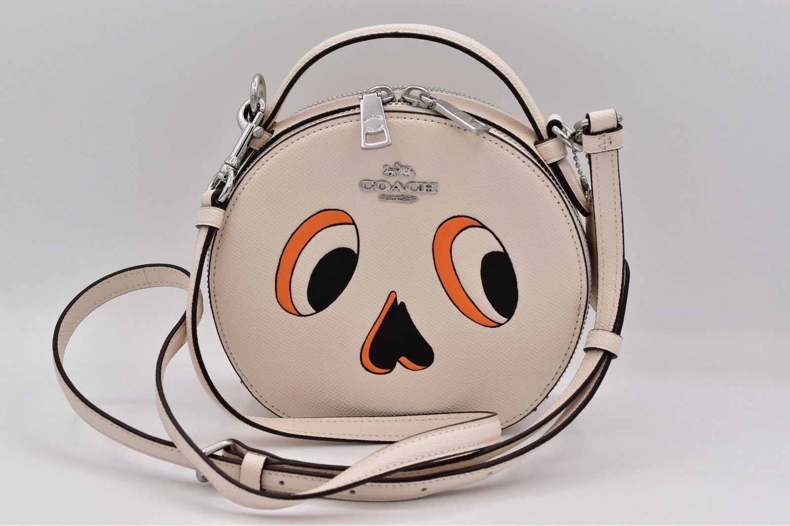 A Coach round shoulder bag in light beige leather with a cartoon face graphic featuring orange and black eyes and a nose, shown on a light gray background.