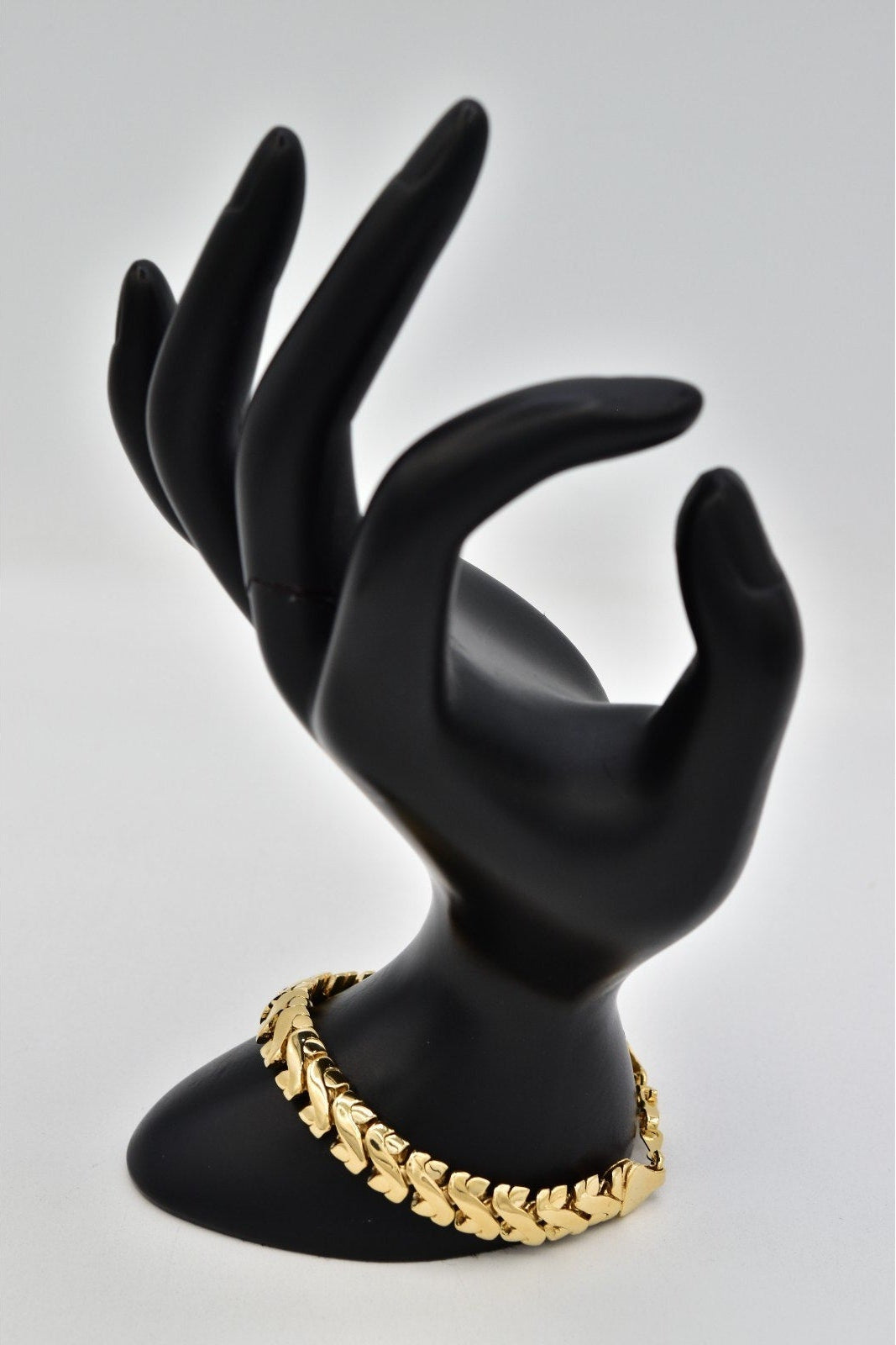 A gold-toned curb link chain bracelet displayed on a matte black hand-shaped jewelry stand against a light gray background.