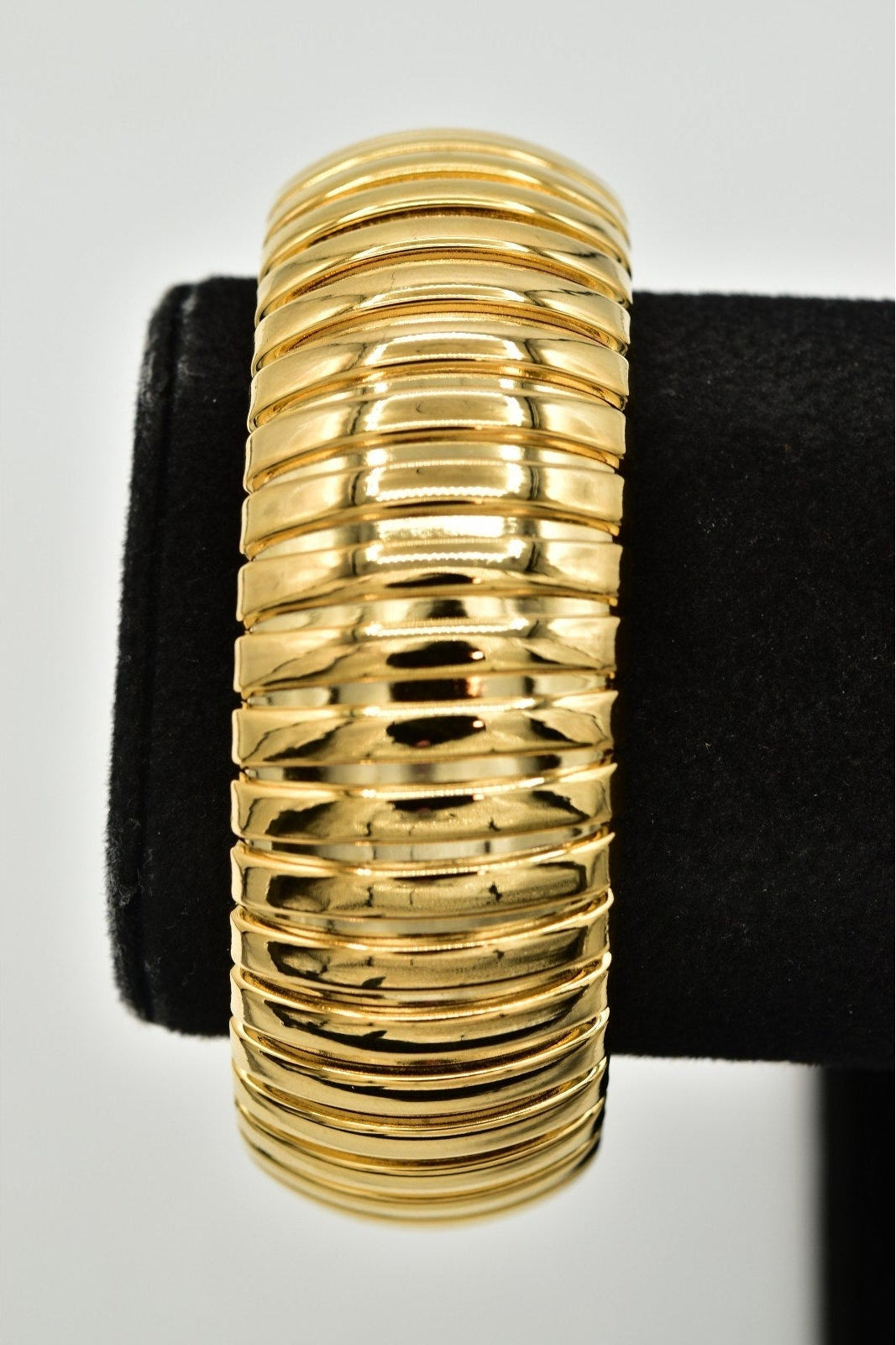 A wide, ribbed gold bangle bracelet displayed on a black velvet ring stand against a light gray background.