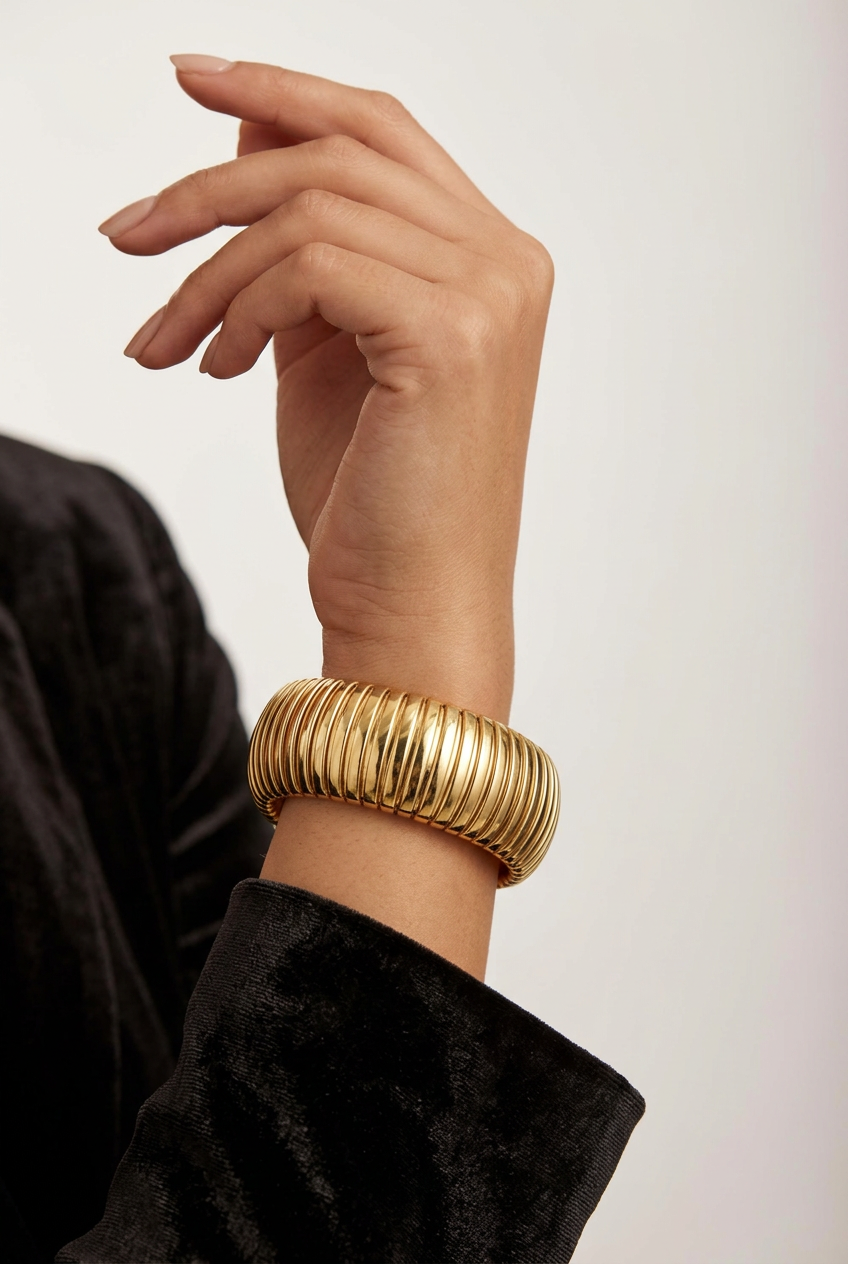 A close-up of a person's hand and wrist wearing a wide, ribbed gold cuff bracelet, paired with a black velvet sleeve, against a plain light background.