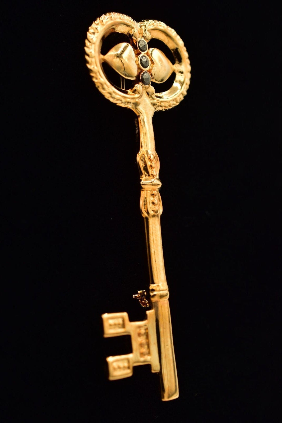 A vintage-style gold key pendant or brooch with ornate detailing, set against a solid black background.