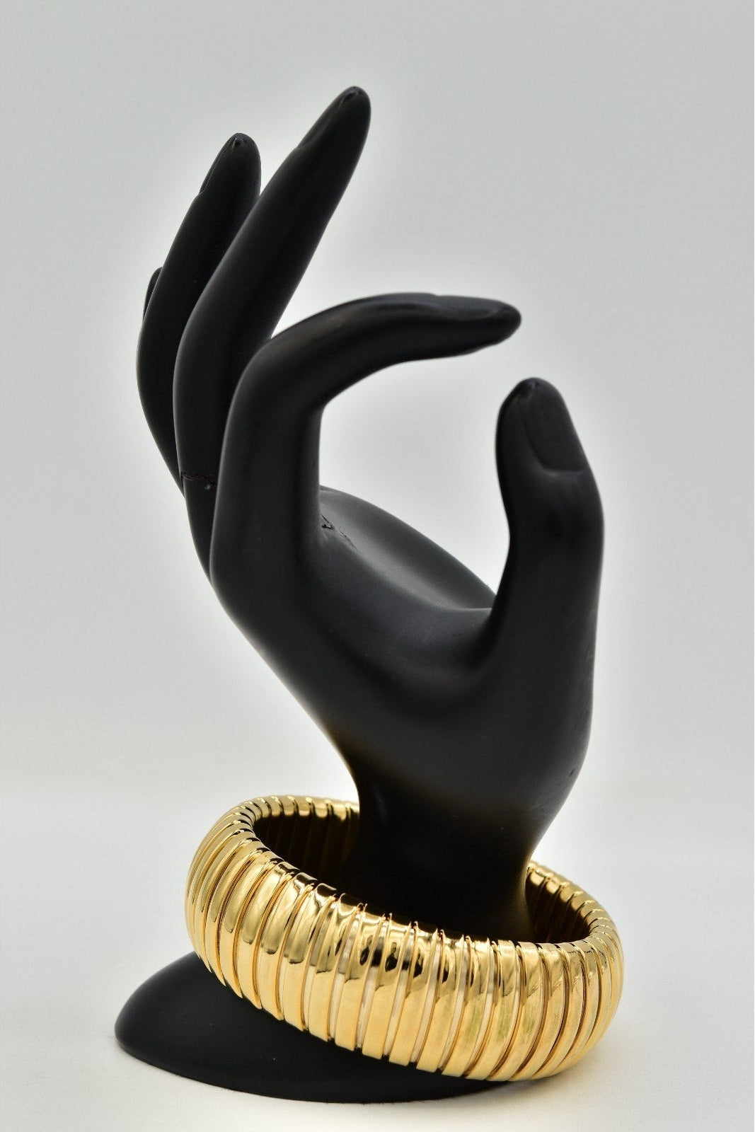 A wide, ribbed gold bangle bracelet displayed on a matte black hand-shaped jewelry stand against a light gray studio background.