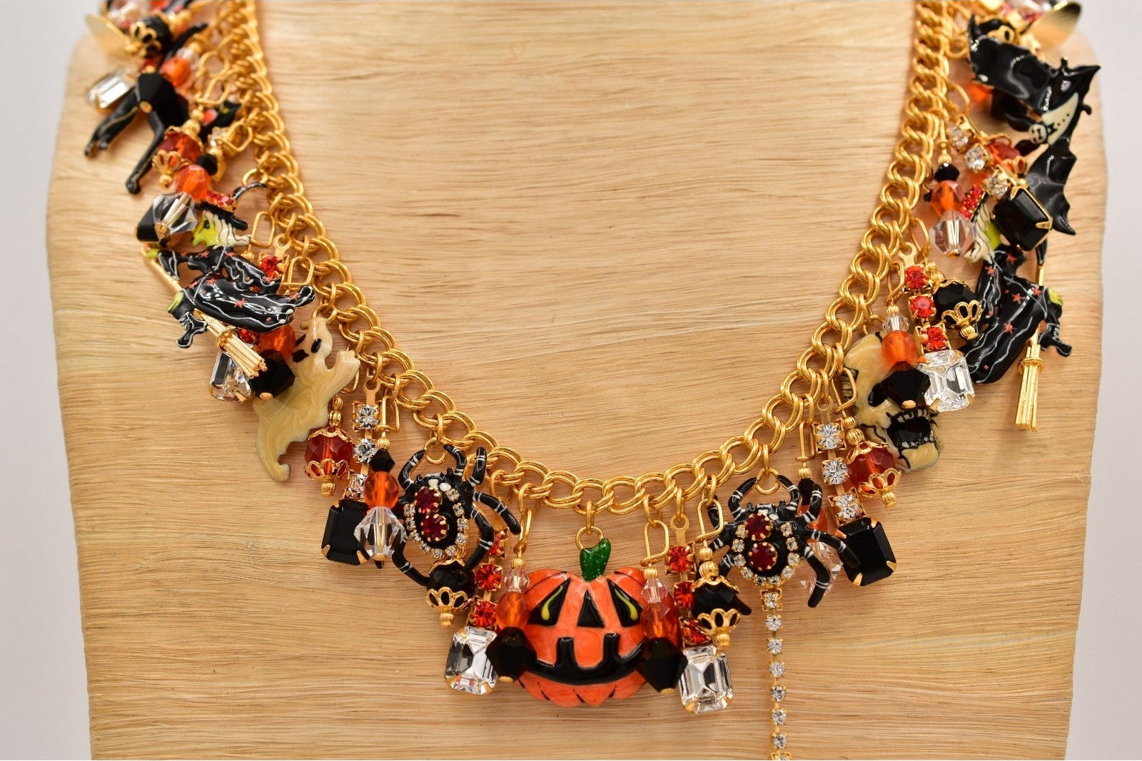 A gold-tone chain necklace with Halloween-themed charms, including a central orange pumpkin, black witch hats, bats, and small decorative figures, displayed on a light wood stand.