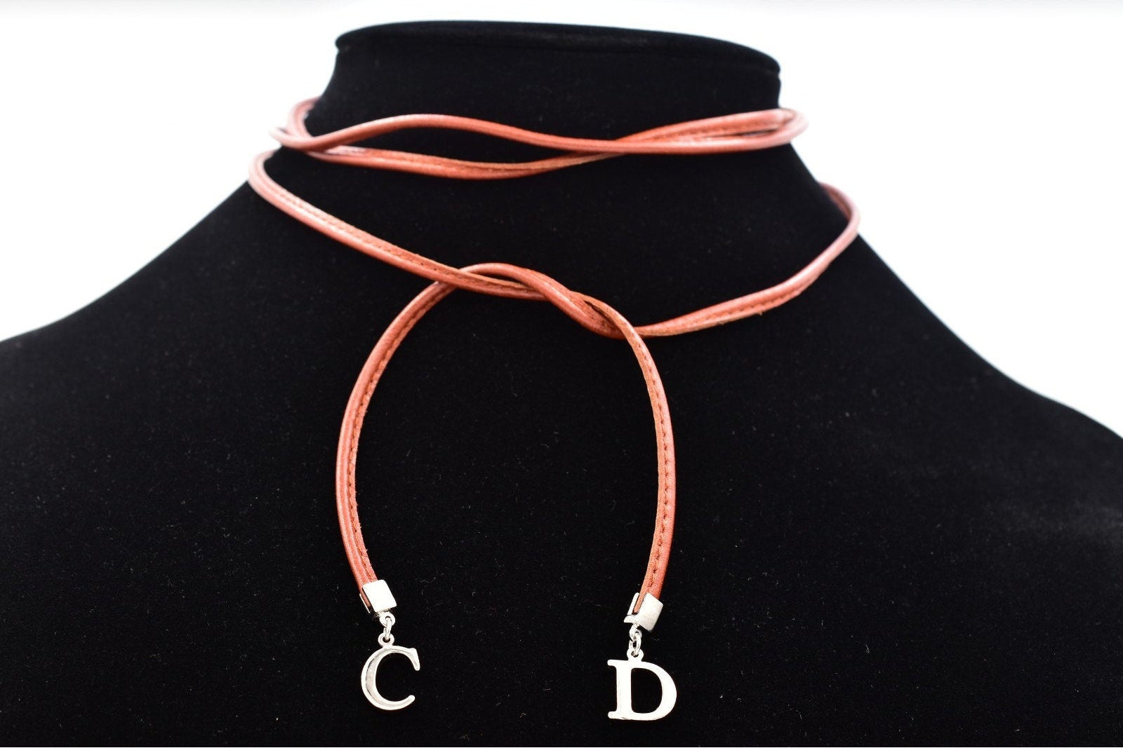 A coral pink faux leather cord necklace with silver letter charms 'C' and 'D', displayed on a black velvet mannequin bust against a white background.