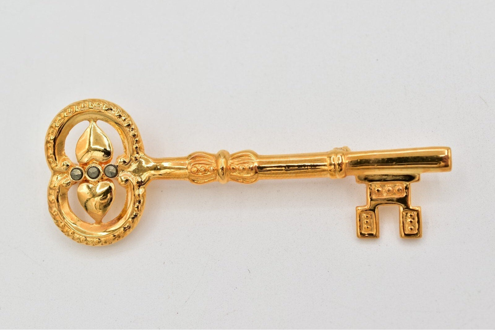 A gold-colored decorative key-shaped brooch or pin with a heart and leaf motif in the wheel, set against a plain light gray background.