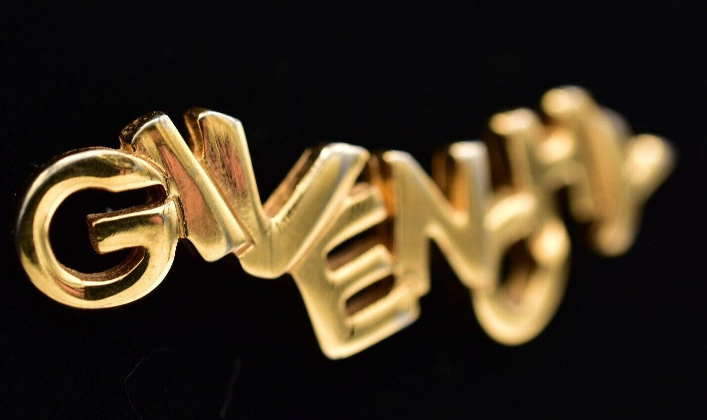 A vintage Givenchy gold logo brooch with block letters on a black background.