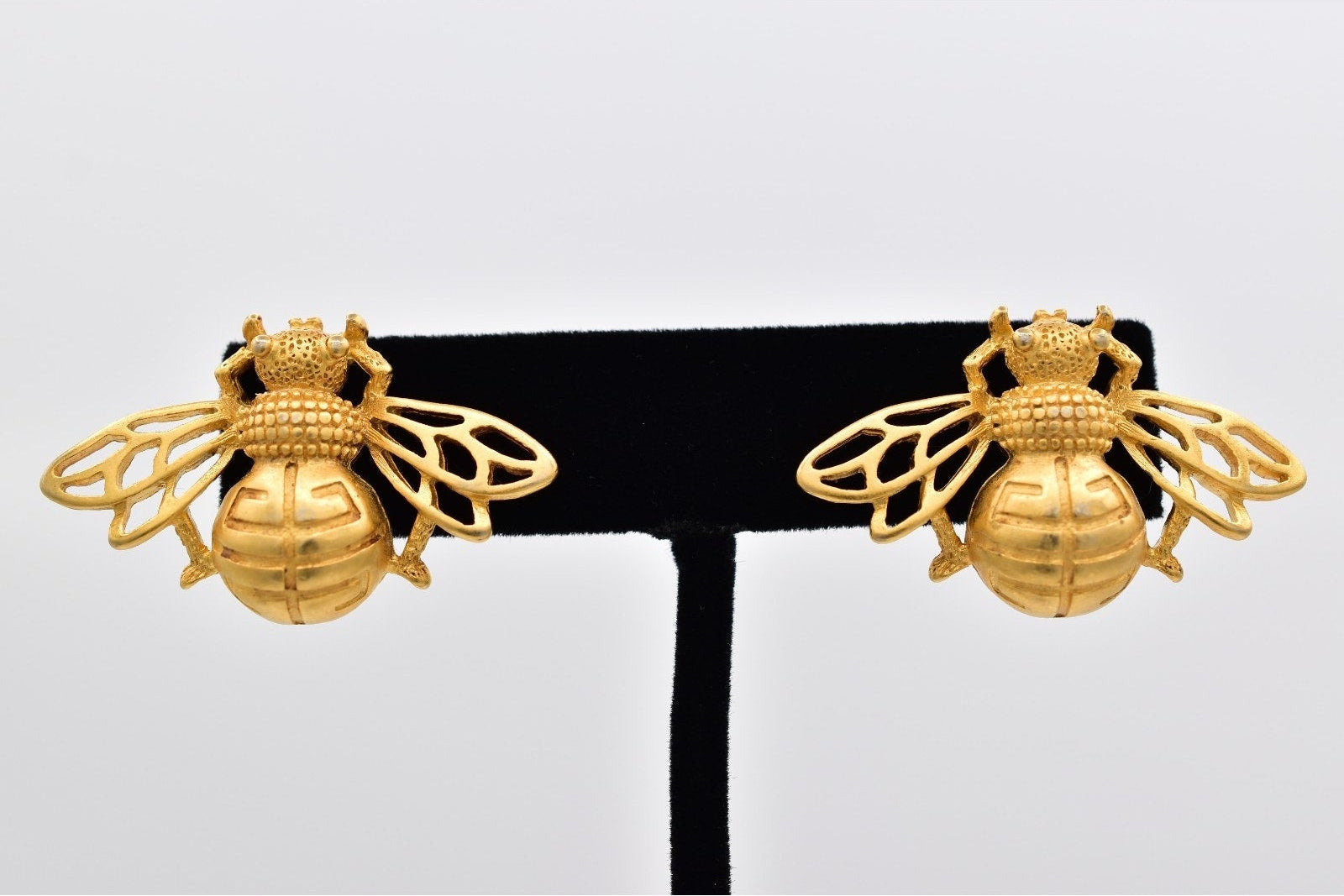 Gold bee-shaped earrings on a black stand against a light gray background