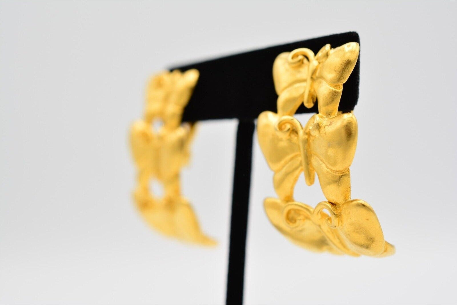 A pair of gold butterfly-shaped earrings displayed on a black stand against a light gray background.