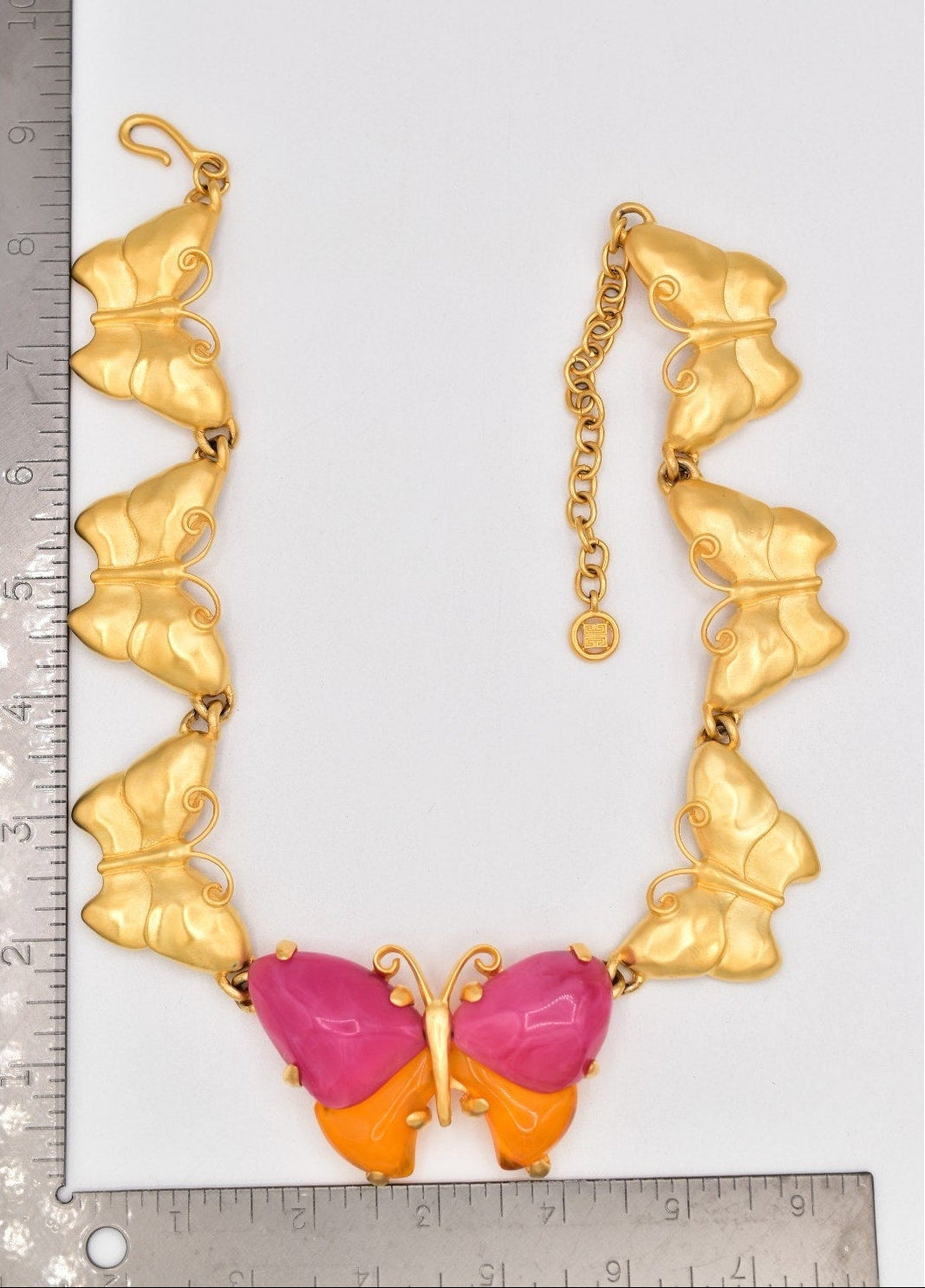 Gold butterfly necklace with pink and orange details next to a ruler for scale on a white background