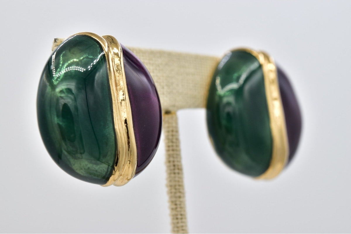 A pair of gold stud earrings with oval green agate and purple amethyst cabochons, displayed on a gold T-stand against a light gray background.