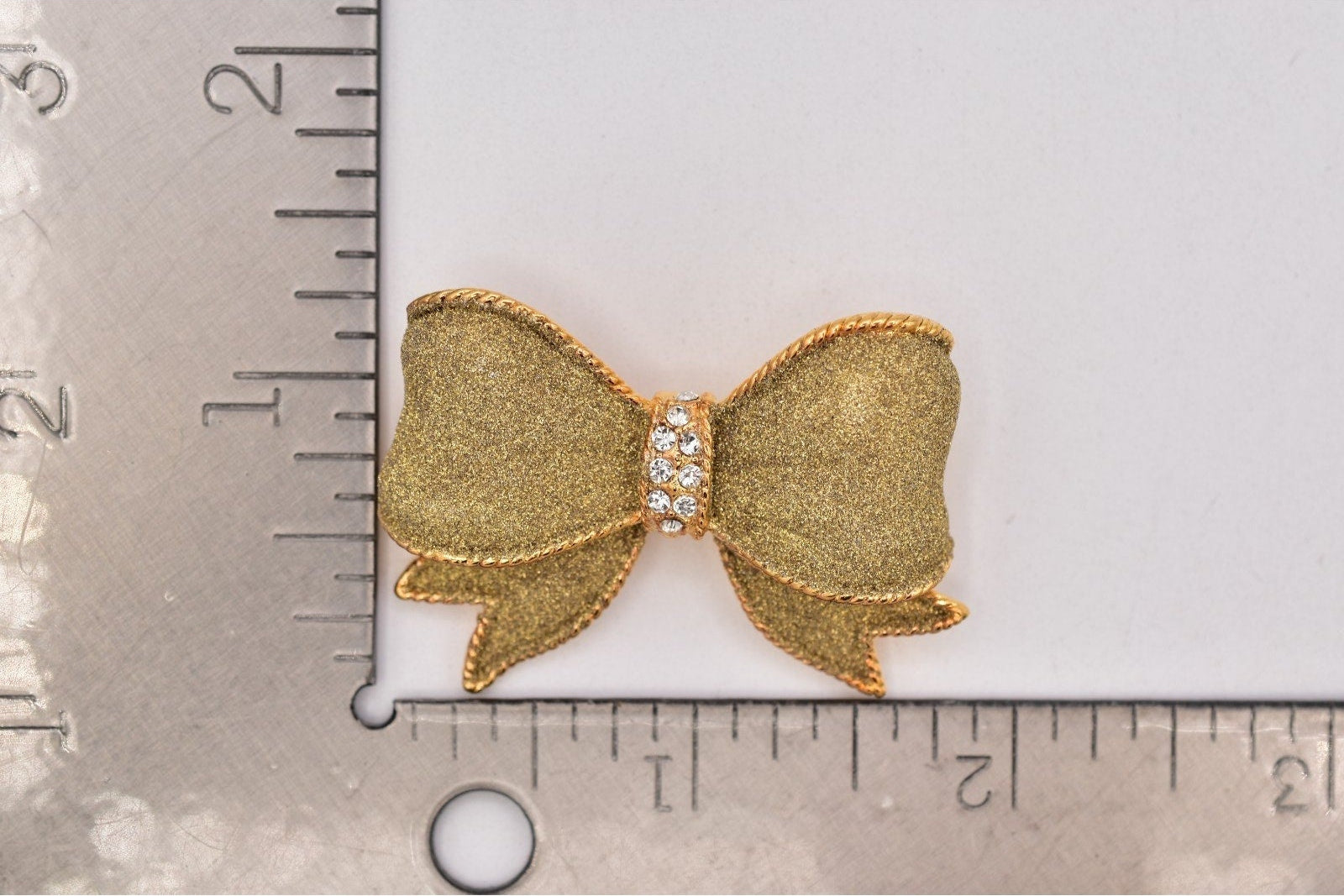 A small gold bow-shaped embellishment or brooch with a rhinestone center, shown on a white background with a metal ruler for scale.