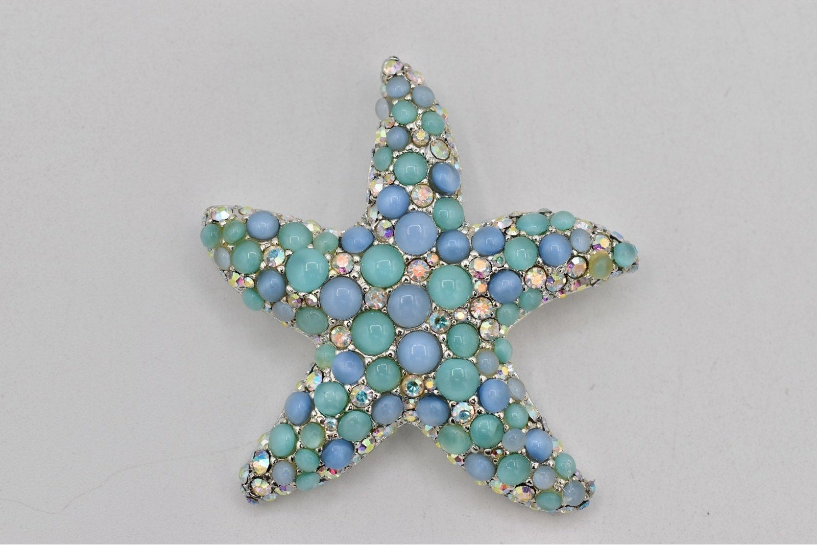 A starfish-shaped brooch embellished with small round blue and mint green rhinestones on a silver-toned setting, shown against a plain light gray background.