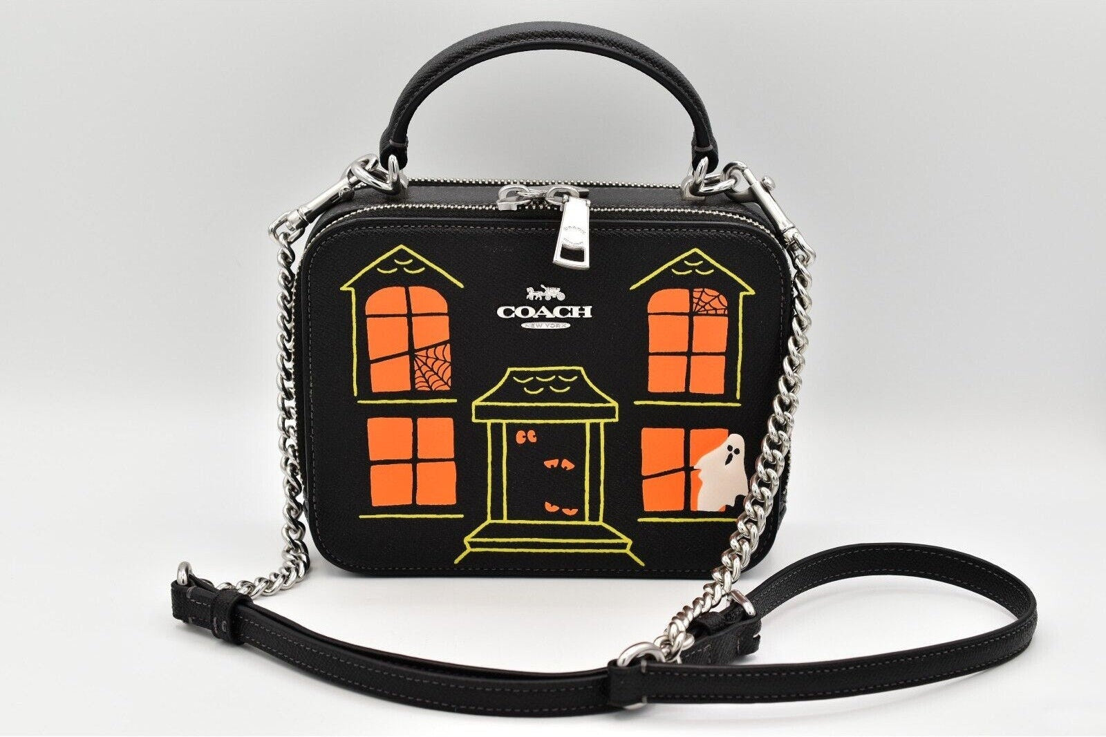 A Coach handbag with a Halloween house design, featuring a black body, orange window panes, yellow trim, a silver chain handle, and a black shoulder strap on a light gray background.