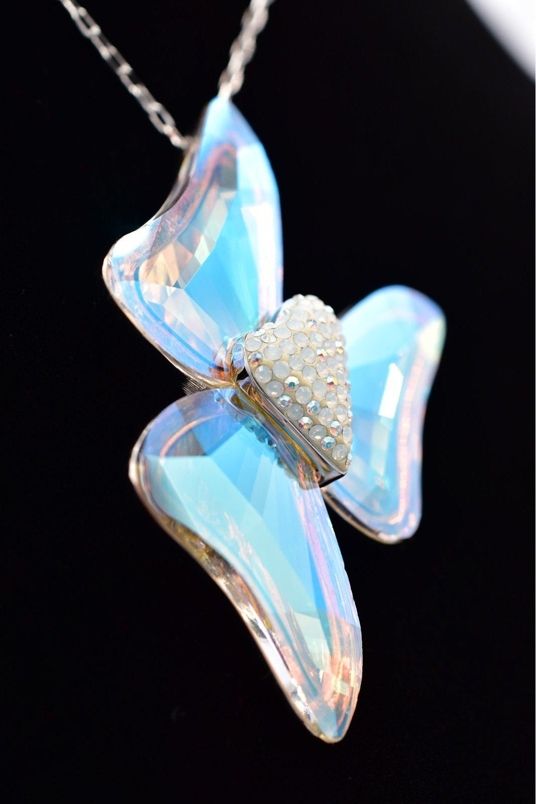 A butterfly-shaped pendant necklace with iridescent light blue and pink opalescent wings, a heart-shaped center paved with small clear stones, and a silver-toned chain against a black background.