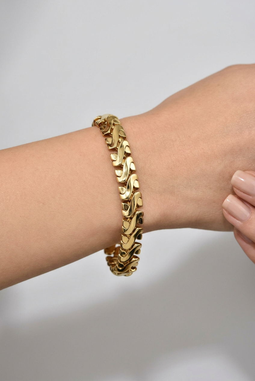 A close-up of a gold chain bracelet with a wavy, textured link design worn on a person's wrist against a neutral light gray background.