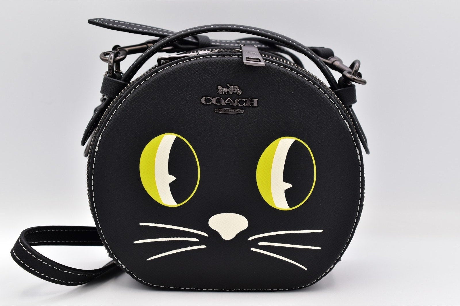 A black Coach crossbody bag shaped like a round cat face, featuring large yellow and white eyes, white whiskers, and a silver-tone COACH logo on the front, shown against a light gray background.
