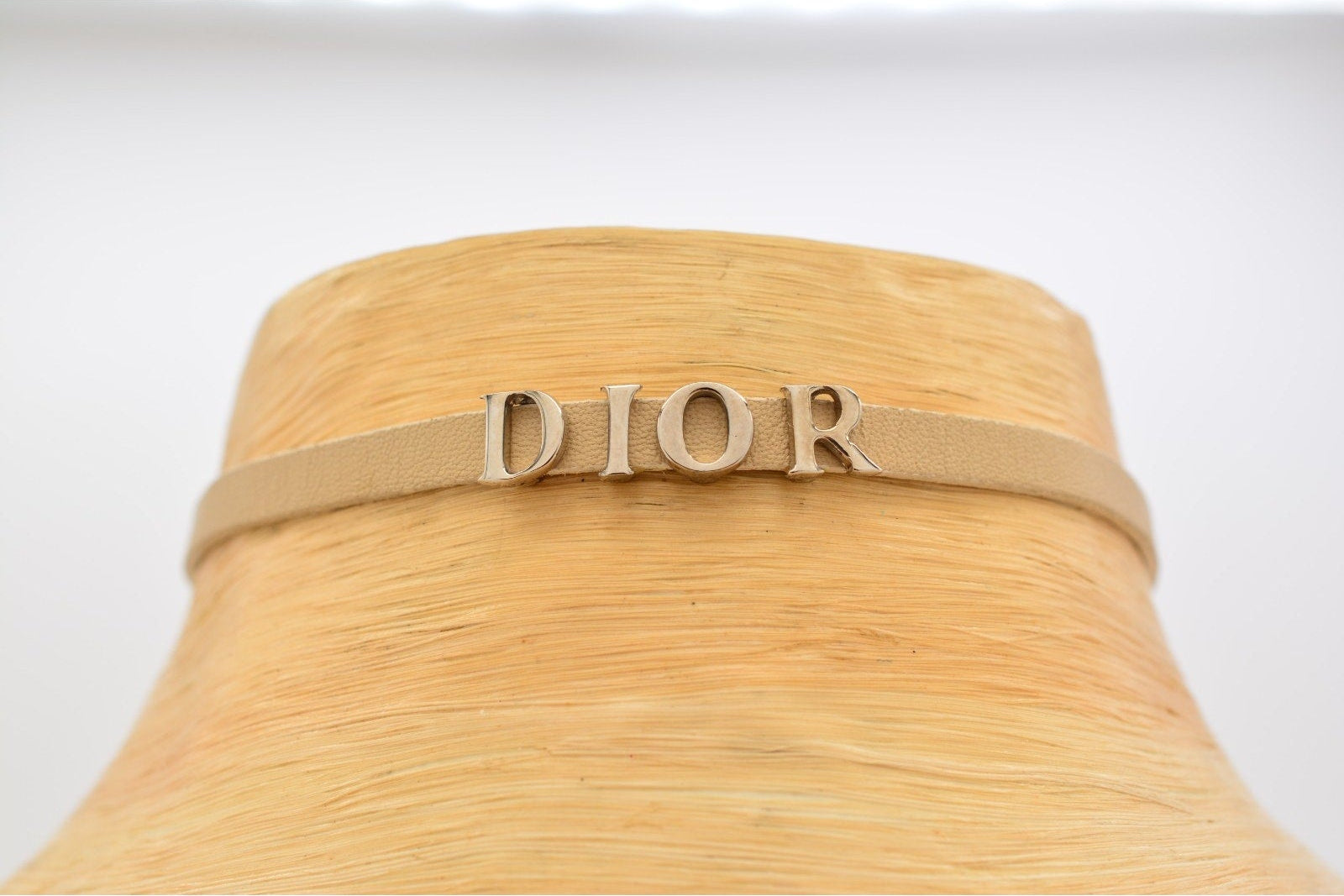 A Dior logo headband in beige faux leather with silver-tone lettering, displayed on a light wood mannequin head against a white background.